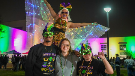 Three students posed at an outdoor event wearing Mardi Gras masks and beads. A stiltwalker poses behind them.